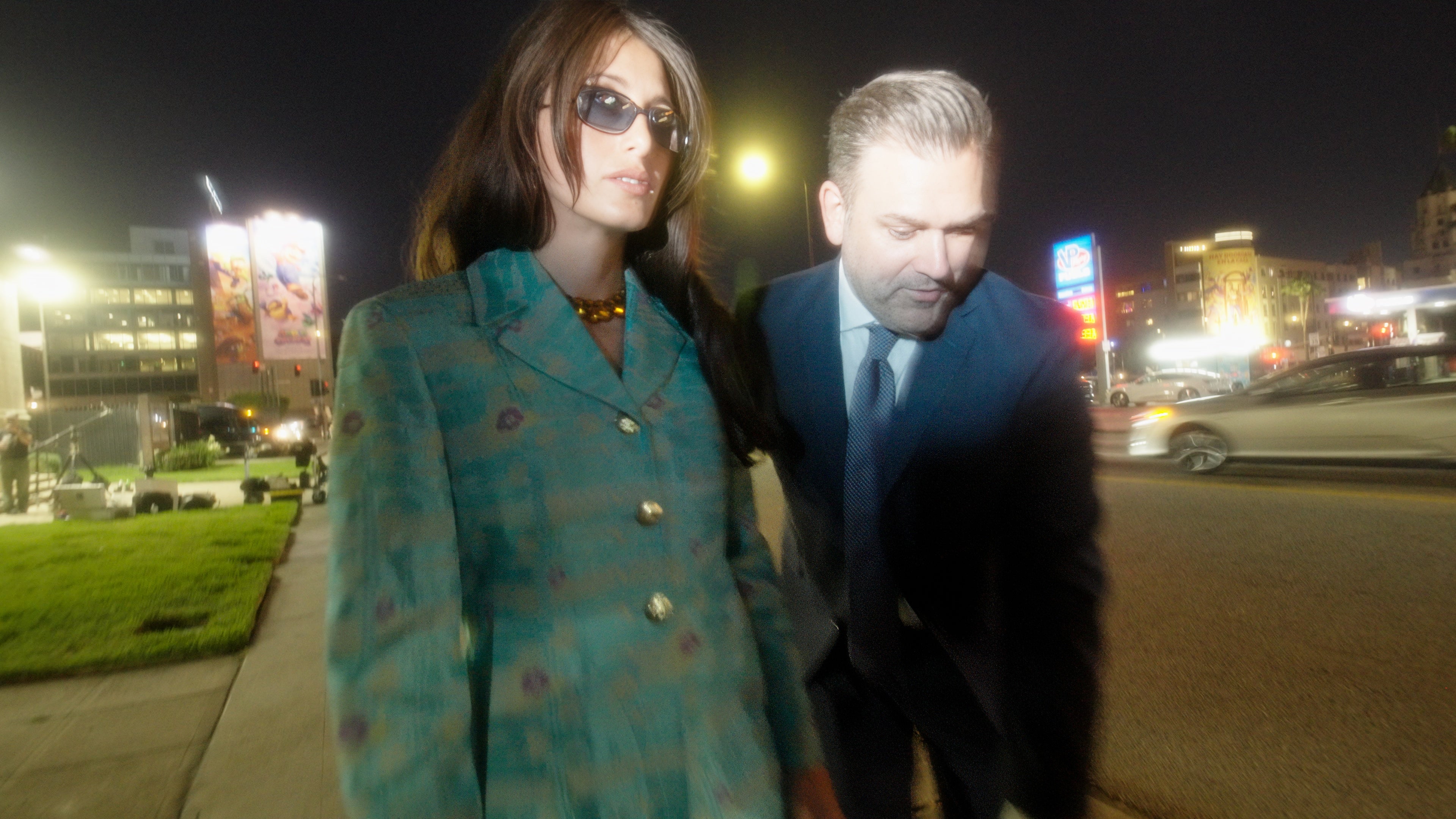 Two people walking on a sidewalk at night with city lights in the background