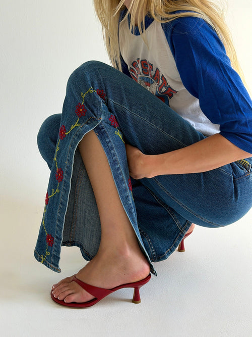 Person wearing blue jeans with floral embroidery and a white shirt with red and black design, sitting on a white background.